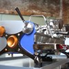 This image is a front-side view of the Slayer Espresso custom machine with custom painted side panels, black nickle plated x-legs, custom painted hubs and accents in Ziricote wood, 3 groups at traditional height with manual dosing controls.